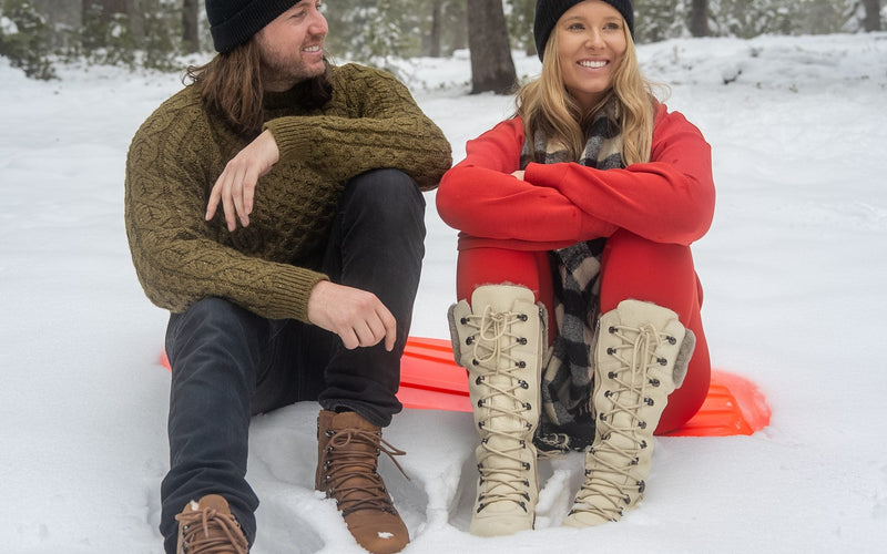 A couple sitting on a sled in the snow, her in tall Pale Ale White DALHOUSIE boots