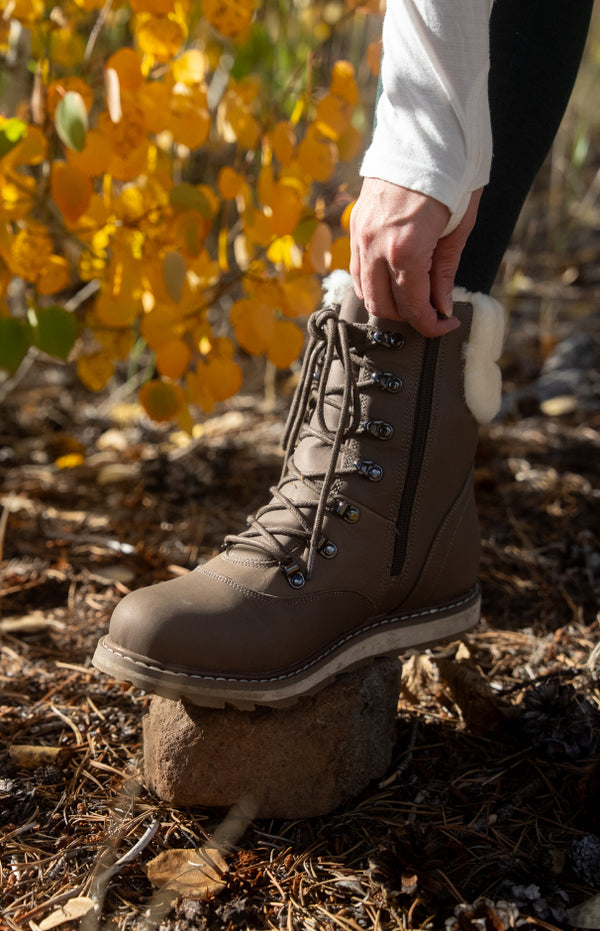 Brown boot with white fur lining worn by a person standing on a rock in a natural setting with yellow leaves.